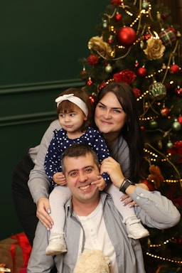 A family poses together in front of a decorated Christmas tree. The father is sitting with his daughter on his shoulders, while the mother stands beside them, smiling. The tree is adorned with baubles, lights, and flowers, creating a festive atmosphere.
