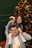 A family poses together in front of a decorated Christmas tree. The father is sitting with his daughter on his shoulders, while the mother stands beside them, smiling. The tree is adorned with baubles, lights, and flowers, creating a festive atmosphere.