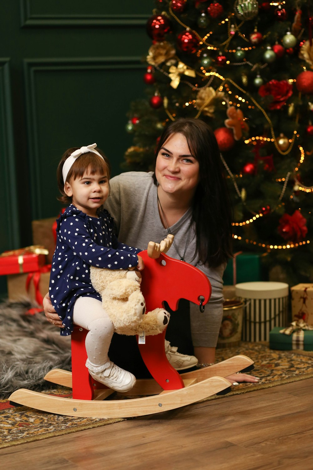 a woman holding a child on a rocking horse in front of a christmas tree
