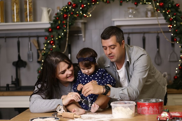 A family is gathered in a kitchen decorated for the holidays. A woman, a child, and a man are enjoying a baking activity. The scene features a wreath adorned with red ornaments in the background and various kitchen utensils hanging from the wall. The countertop is cluttered with a rolling pin, a container of flour, and a festive tin.