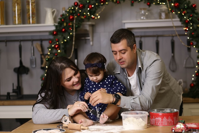 A family is gathered in a kitchen decorated for the holidays. A woman, a child, and a man are enjoying a baking activity. The scene features a wreath adorned with red ornaments in the background and various kitchen utensils hanging from the wall. The countertop is cluttered with a rolling pin, a container of flour, and a festive tin.
