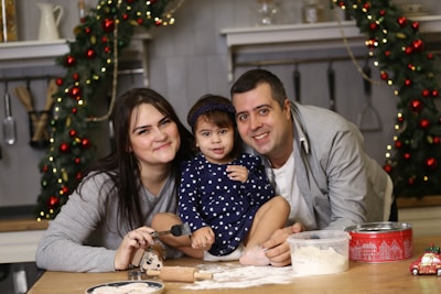 A family of three is gathered around a table in a festive kitchen. The young girl in the center is sitting on the table wearing a polka dot dress, surrounded by her parents. Holiday decorations such as garlands with red baubles adorn the background, and there are baking items like dough, flour, and rolling pin present.