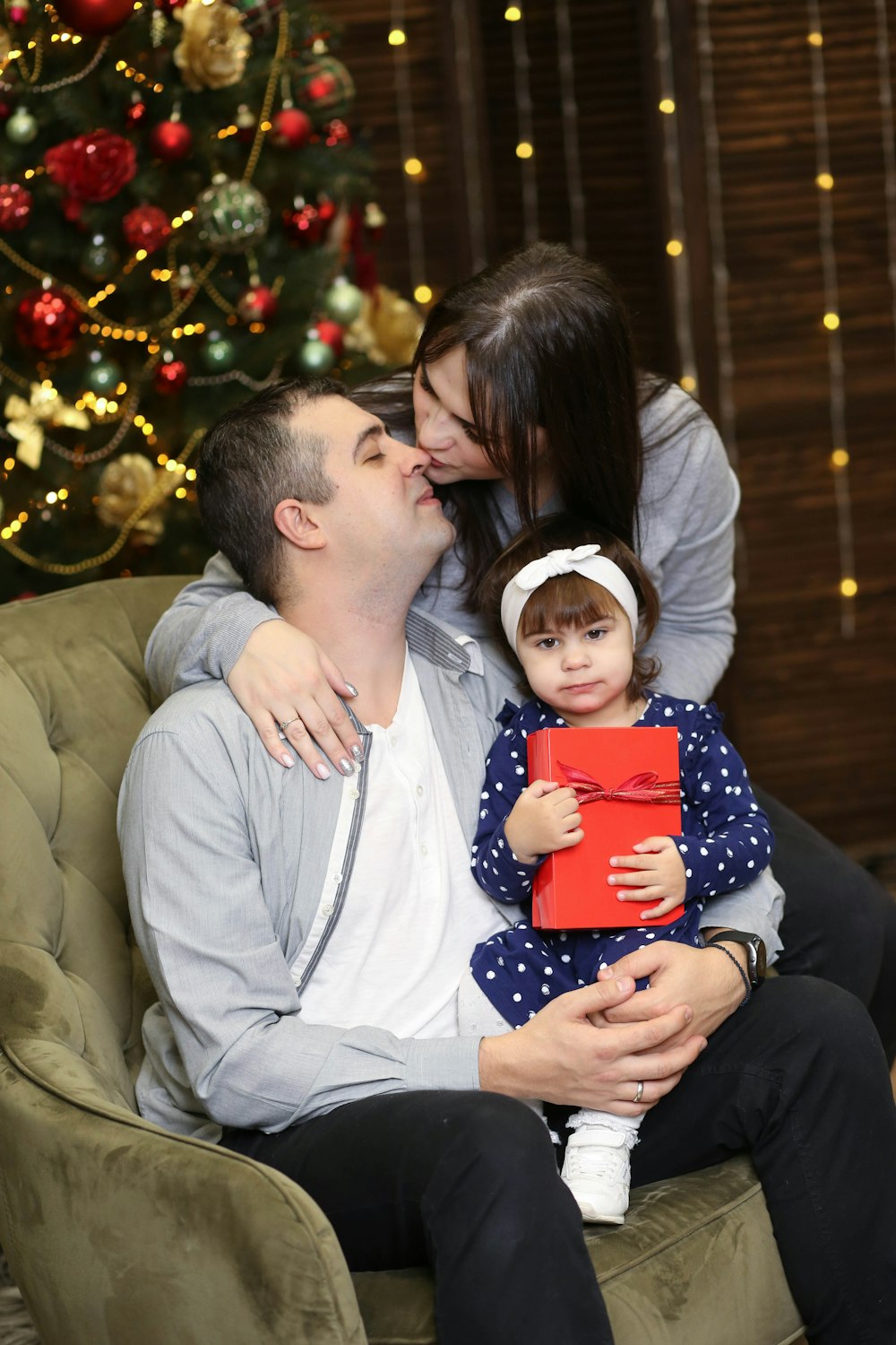 a man, woman and child sitting on a chair in front of a christmas tree