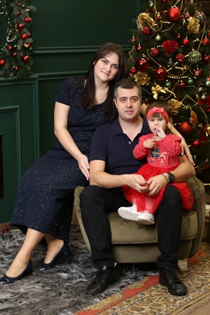 A family of three is seated in a cozy room with Christmas decorations, including a decorated tree with red and gold ornaments. The adults, a man and a woman, are sitting on a plush green chair, and they are holding a child dressed in a red outfit with a headband. The room has a warm, festive atmosphere.
