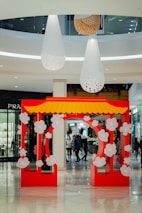 A vibrant red and yellow decorative archway adorned with white floral patterns stands prominently in the foreground of a shopping mall. Large white and wooden hanging fixtures cascade from the ceiling above, adding a modern touch to the space. Shoppers can be seen walking in the background amidst high-end store displays.