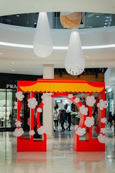 A vibrant red and yellow decorative archway adorned with white floral patterns stands prominently in the foreground of a shopping mall. Large white and wooden hanging fixtures cascade from the ceiling above, adding a modern touch to the space. Shoppers can be seen walking in the background amidst high-end store displays.