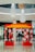 A vibrant red and yellow decorative archway adorned with white floral patterns stands prominently in the foreground of a shopping mall. Large white and wooden hanging fixtures cascade from the ceiling above, adding a modern touch to the space. Shoppers can be seen walking in the background amidst high-end store displays.