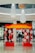 A vibrant red and yellow decorative archway adorned with white floral patterns stands prominently in the foreground of a shopping mall. Large white and wooden hanging fixtures cascade from the ceiling above, adding a modern touch to the space. Shoppers can be seen walking in the background amidst high-end store displays.