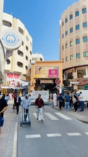 A bustling street scene with people walking and shopping in an urban area. There are multiple buildings with signs, including a prominent sign for 'Main Point' and another advertising 'Malabar Gold & Diamonds'. A man is pushing a cart while another person walks alongside. The setting includes a mix of modern and traditional architecture.