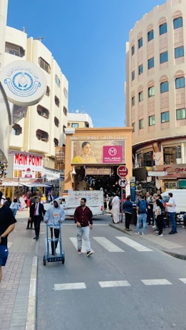 A bustling street scene with people walking and shopping in an urban area. There are multiple buildings with signs, including a prominent sign for 'Main Point' and another advertising 'Malabar Gold & Diamonds'. A man is pushing a cart while another person walks alongside. The setting includes a mix of modern and traditional architecture.