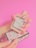 A hand with long, manicured nails holds a rectangular perfume bottle with a minimalist design. The label on the bottle reads 'Gucci Bloom'. Below, another hand supports an ornate box with a floral pattern. The background is bright pink.