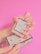 A hand with long, manicured nails holds a rectangular perfume bottle with a minimalist design. The label on the bottle reads 'Gucci Bloom'. Below, another hand supports an ornate box with a floral pattern. The background is bright pink.