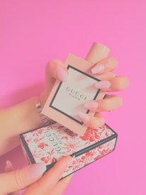 A hand with long, manicured nails holds a rectangular perfume bottle with a minimalist design. The label on the bottle reads 'Gucci Bloom'. Below, another hand supports an ornate box with a floral pattern. The background is bright pink.