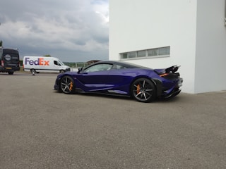 A sleek, dark blue sports car with an aerodynamic design is parked near a white building. The car's orange brake calipers are visible through its multi-spoke wheels. In the background, a FedEx delivery van is parked beside a black van, with a cloudy sky overhead.