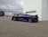A sleek, dark blue sports car with an aerodynamic design is parked near a white building. The car's orange brake calipers are visible through its multi-spoke wheels. In the background, a FedEx delivery van is parked beside a black van, with a cloudy sky overhead.