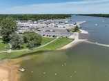 A vibrant marina area with numerous parked boats and vehicles. The scene includes a body of water with boats sailing and a beach with people enjoying leisure activities. Greenery surrounds the area, and there is a large parking lot filled with cars.