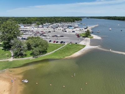 A vibrant marina area with numerous parked boats and vehicles. The scene includes a body of water with boats sailing and a beach with people enjoying leisure activities. Greenery surrounds the area, and there is a large parking lot filled with cars.