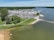 A vibrant marina area with numerous parked boats and vehicles. The scene includes a body of water with boats sailing and a beach with people enjoying leisure activities. Greenery surrounds the area, and there is a large parking lot filled with cars.