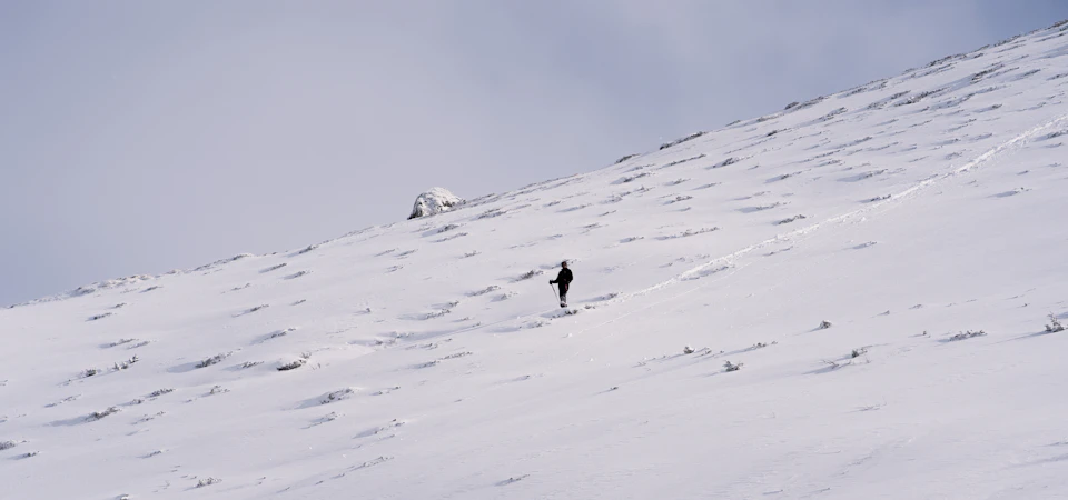 The image depicts a lone skier descending a snowy mountain slope, wearing a black jacket and pants against the white snow. The skier is moving towards the right side of the frame, leaving behind a trail on the pristine white snow. In the distance, there are rocks visible in the background, adding to the rugged beauty of the scene.