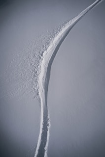 A single, curved trail is carved through an expanse of smooth, undisturbed snow, suggesting a path made by a skier or snowboarder. The surrounding snow shows subtle texture, with shadows adding depth to the scene.