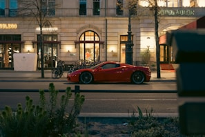 A sleek red sports car is parked on a city street in front of a high-end hotel. The street is lined with bicycles, and the hotel features grand architectural elements with well-lit entrances and decorative windows. The setting is urban, with nearby trees and plants visible in the foreground.