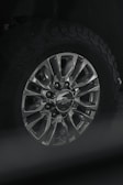 A close-up view of a car wheel with a metallic alloy rim featuring multiple spokes and a center cap displaying a logo. The tire attached is rugged with a distinct tread pattern, likely suitable for off-road driving.