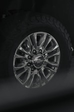 A close-up view of a car wheel with a metallic alloy rim featuring multiple spokes and a center cap displaying a logo. The tire attached is rugged with a distinct tread pattern, likely suitable for off-road driving.