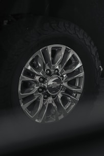 A close-up view of a car wheel with a metallic alloy rim featuring multiple spokes and a center cap displaying a logo. The tire attached is rugged with a distinct tread pattern, likely suitable for off-road driving.