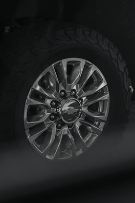 A close-up view of a car wheel with a metallic alloy rim featuring multiple spokes and a center cap displaying a logo. The tire attached is rugged with a distinct tread pattern, likely suitable for off-road driving.