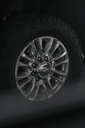 A close-up view of a car wheel with a metallic alloy rim featuring multiple spokes and a center cap displaying a logo. The tire attached is rugged with a distinct tread pattern, likely suitable for off-road driving.