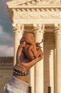 A hand is holding a small doll resembling a newborn baby. In the background, there is a large neoclassical building with columns, possibly a government or courthouse structure. The hand is adorned with multiple bracelets.