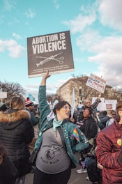 A group of people is gathered in a public area, participating in a protest. A central figure, wearing a jacket with a rainbow patch and a 'Human Rights Start Here' shirt, holds a large sign reading 'Abortion is Violence'. Other individuals hold various signs and banners. The setting appears to be an urban environment with city buildings, under a clear blue sky with clouds.