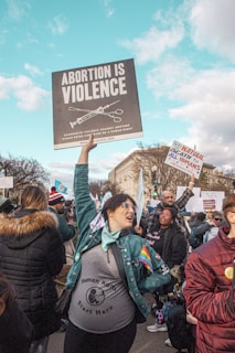A group of people is gathered in a public area, participating in a protest. A central figure, wearing a jacket with a rainbow patch and a 'Human Rights Start Here' shirt, holds a large sign reading 'Abortion is Violence'. Other individuals hold various signs and banners. The setting appears to be an urban environment with city buildings, under a clear blue sky with clouds.