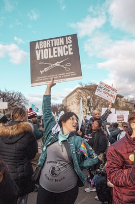 A group of people is gathered in a public area, participating in a protest. A central figure, wearing a jacket with a rainbow patch and a 'Human Rights Start Here' shirt, holds a large sign reading 'Abortion is Violence'. Other individuals hold various signs and banners. The setting appears to be an urban environment with city buildings, under a clear blue sky with clouds.