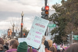 A protest rally with a crowd of people holding signs, one prominently displaying a message about abortion. The setting appears to be outdoors on a street with traffic lights and trees in the background.