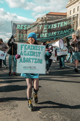 A person at a protest holds a sign reading 'Feminists Against Abortion' while wearing a blue hat and skirt, surrounded by others carrying various banners. The main banner in the background reads 'Respect Existence or Expect Resistance.'