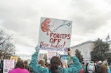 A group of people participating in a protest or march. One person in the foreground is holding a large sign with an image of a fetus and a pair of forceps, alongside the text 'FORCEPS OFF their bodies! RehumanizeIntl'. Other signs in the background include phrases like 'STOP CALLING VIOLENCE FEMINISM'. The participants are surrounded by trees and a building with an American flag.