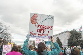 A group of people participating in a protest or march. One person in the foreground is holding a large sign with an image of a fetus and a pair of forceps, alongside the text 'FORCEPS OFF their bodies! RehumanizeIntl'. Other signs in the background include phrases like 'STOP CALLING VIOLENCE FEMINISM'. The participants are surrounded by trees and a building with an American flag.