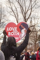 A person holds up a heart-shaped sign displaying the message 'Love Them Both' in a crowded outdoor setting. The sign features an illustration of a woman holding a child. The background includes leafless trees and a building with a spire.