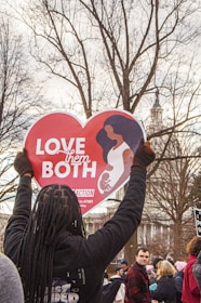 A person holds up a heart-shaped sign displaying the message 'Love Them Both' in a crowded outdoor setting. The sign features an illustration of a woman holding a child. The background includes leafless trees and a building with a spire.