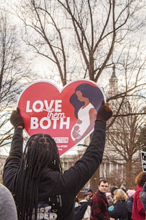 A person holds up a heart-shaped sign displaying the message 'Love Them Both' in a crowded outdoor setting. The sign features an illustration of a woman holding a child. The background includes leafless trees and a building with a spire.