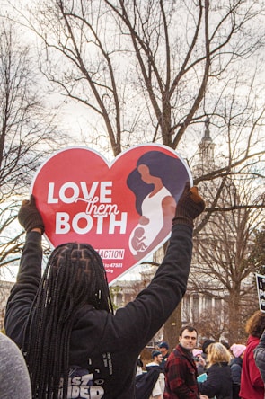 A person holds up a heart-shaped sign displaying the message 'Love Them Both' in a crowded outdoor setting. The sign features an illustration of a woman holding a child. The background includes leafless trees and a building with a spire.