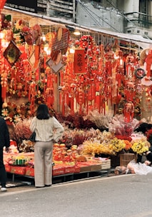 A bustling market stall is adorned with numerous red decorations, lanterns, and hanging ornaments. Flowers and other decorative items are displayed, adding vibrant colors to the scene. A person, wearing a cream sweater and beige pants, stands in front, seemingly browsing or deciding on a purchase.