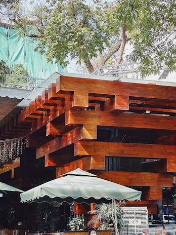 A modern architectural structure featuring interlocking wooden beams with a polished surface. The building is surrounded by lush green trees and has outdoor umbrellas for shade, suggesting an area for seating or a café. There are people in the scene, and some potted plants add a touch of nature to the setting.