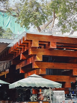 A modern architectural structure featuring interlocking wooden beams with a polished surface. The building is surrounded by lush green trees and has outdoor umbrellas for shade, suggesting an area for seating or a café. There are people in the scene, and some potted plants add a touch of nature to the setting.
