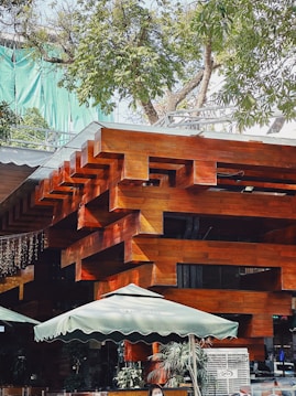 A modern architectural structure featuring interlocking wooden beams with a polished surface. The building is surrounded by lush green trees and has outdoor umbrellas for shade, suggesting an area for seating or a caf&eacute;. There are people in the scene, and some potted plants add a touch of nature to the setting.