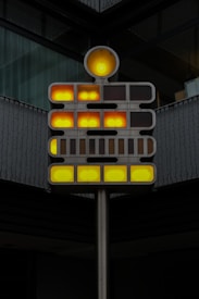 A modern art installation composed of a metallic structure with illuminated circular and rectangular lights. The lights are arranged in a grid pattern, displaying shades of yellow and orange. The installation is situated against a backdrop of a building with dark-colored walls and reflective glass.