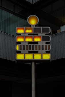 A modern art installation composed of a metallic structure with illuminated circular and rectangular lights. The lights are arranged in a grid pattern, displaying shades of yellow and orange. The installation is situated against a backdrop of a building with dark-colored walls and reflective glass.