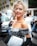 A fashionable person with styled blonde hair and bright red lipstick, wearing an off-the-shoulder black and white printed dress and holding a white designer clutch bag. The setting is urban, with other people in casual and business attire in the background and a parked delivery van nearby.