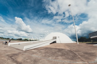 A large, modern white dome building with a long ramp leading up to its entrance is situated in an open area. The sky is partly cloudy with patches of blue showing through. Surrounding the building, there are some trees and distant buildings, along with a tall light pole. The ground appears paved and the scene has an overall expansive feeling.